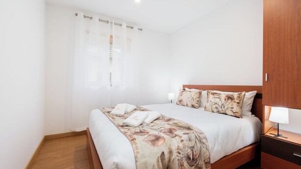 Ein ordentlich bezogenes Bett mit floralem Überwurf, ein Nachttisch aus Holz mit einer Lampe und ein Holzboden.