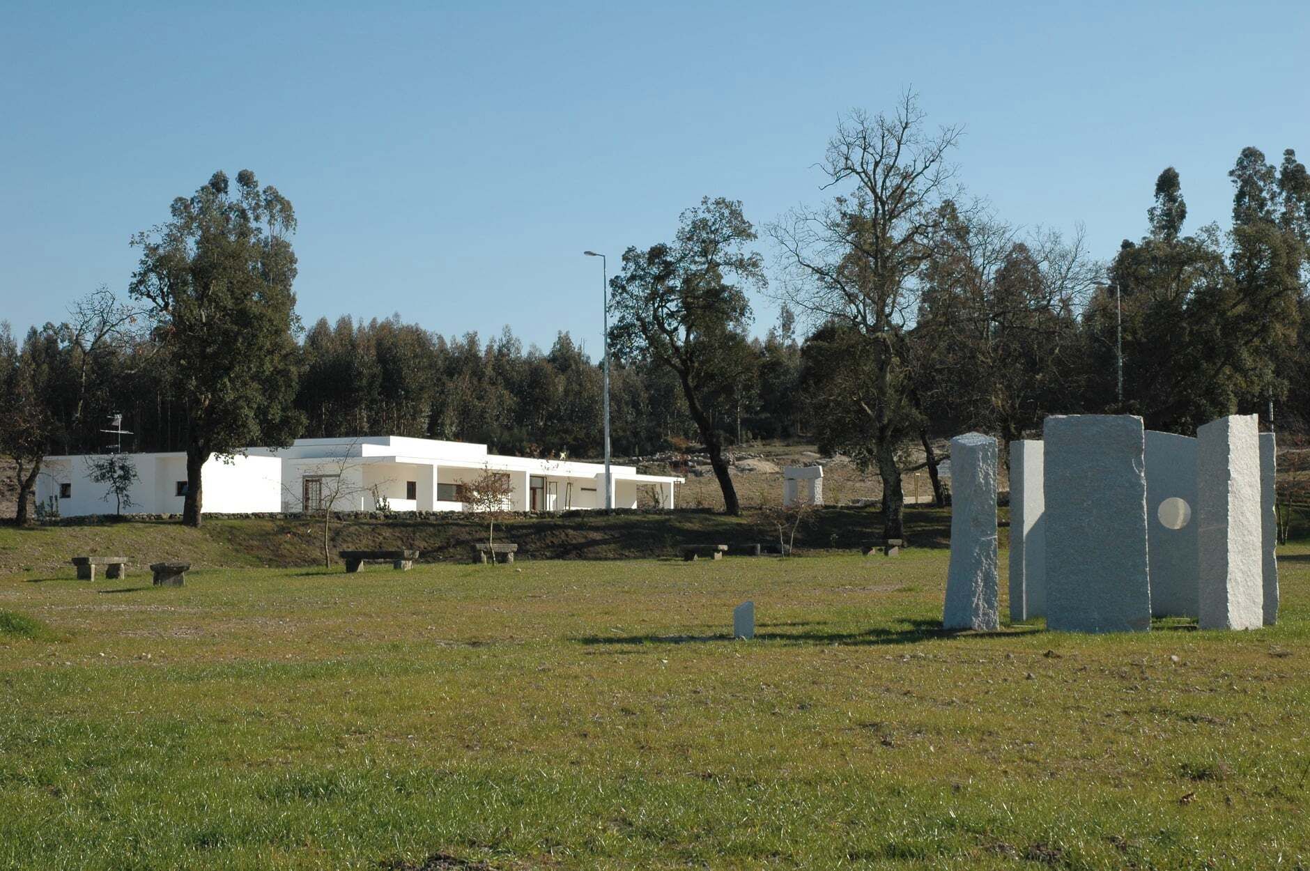 A modern building with a flat roof and white walls, surrounded by a grassy field and trees.