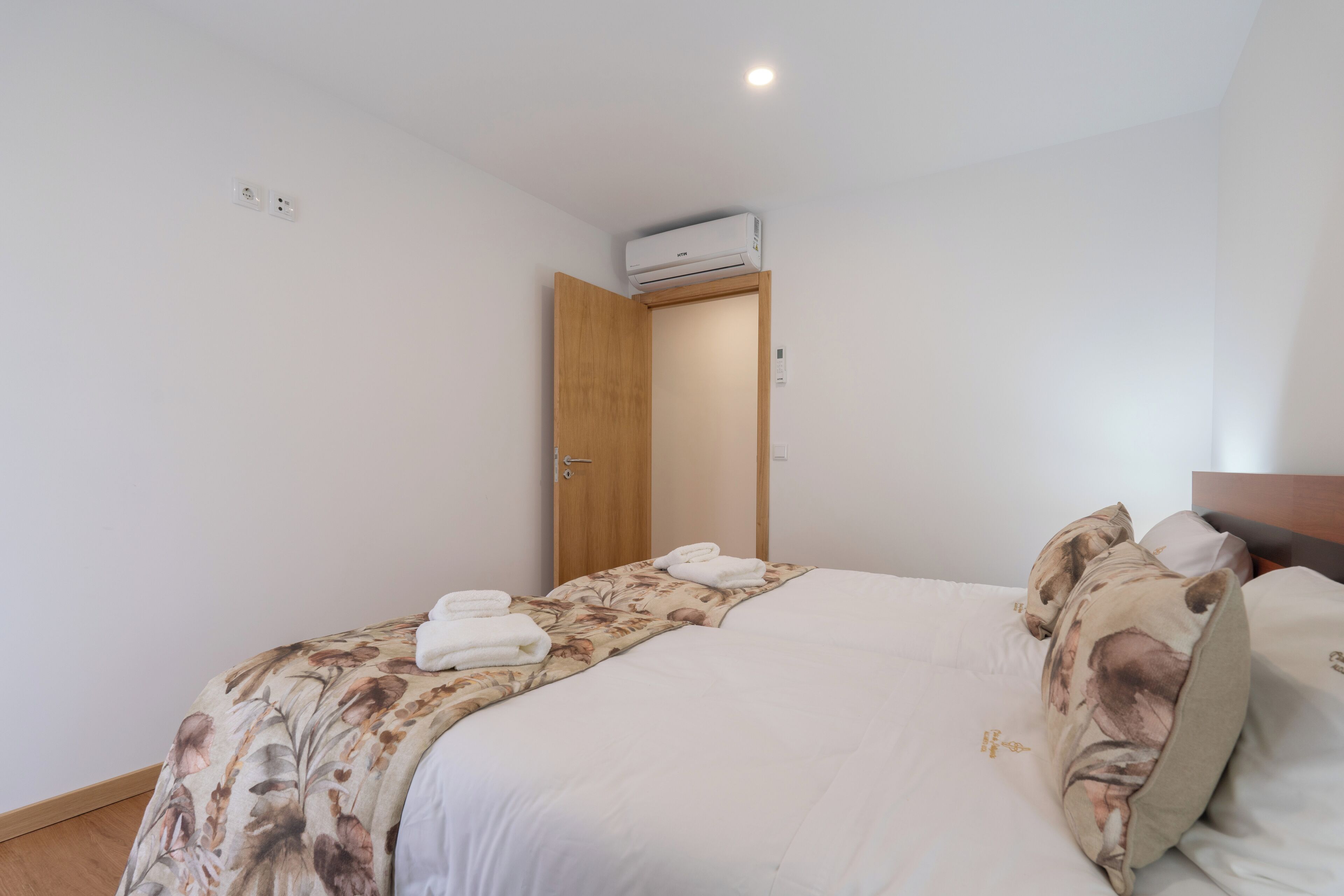 Una cama bien hecha con ropa de cama floral y una colcha blanca, una puerta de madera y un equipo de aire acondicionado montado en la pared.