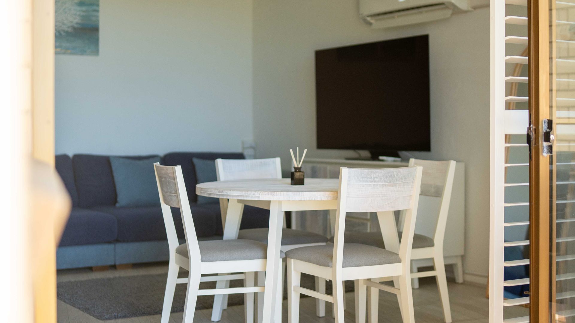 A dining area with a round table and four chairs, a dark-colored sofa, and a flat-screen TV mounted on the wall.