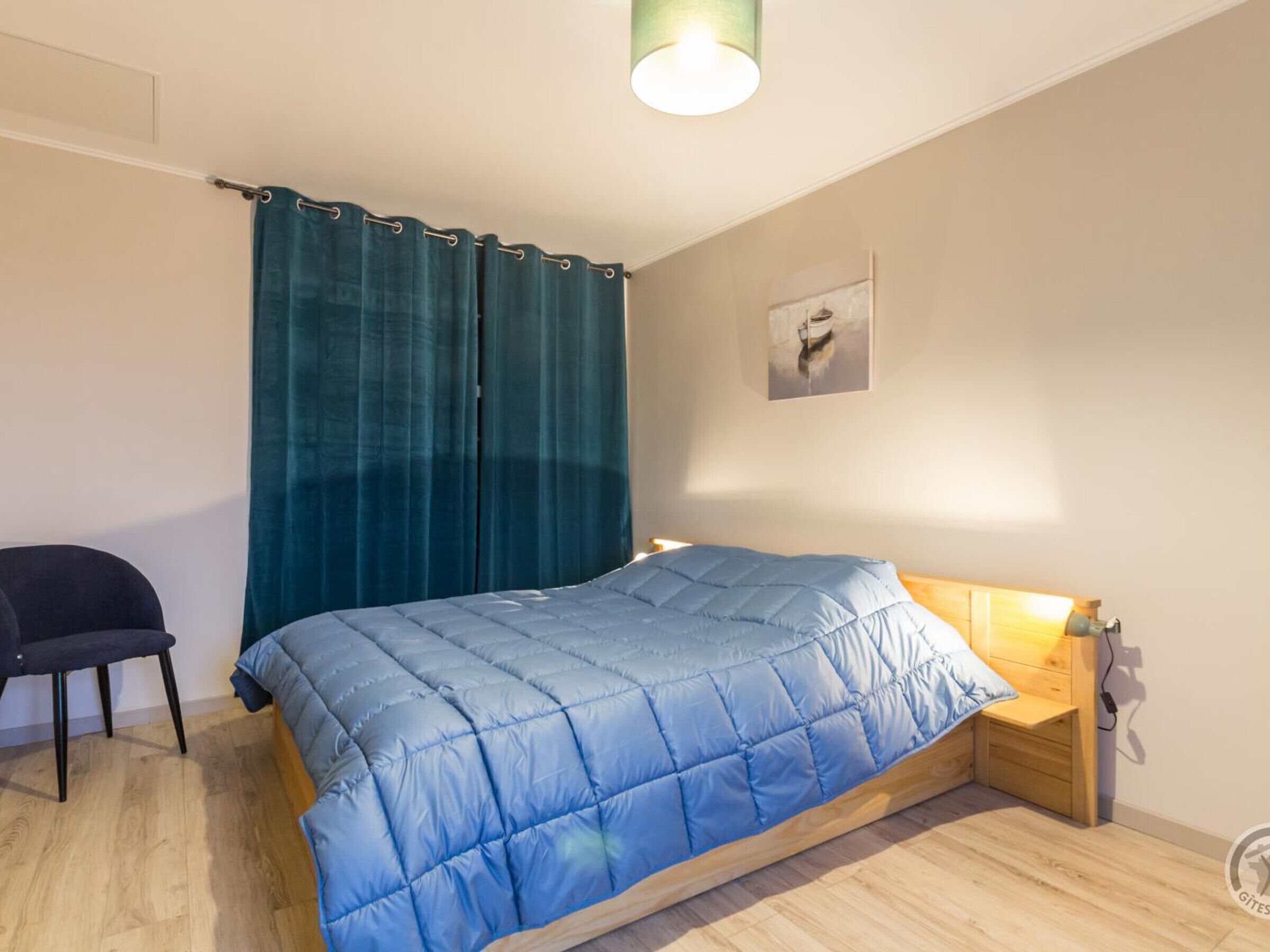Een slaapkamer met een bed, een stoel, een houten nachtkastje, een wandlamp en een schilderij.