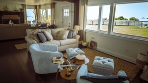 Un salon spacieux doté d’une grande fenêtre donnant sur l’extérieur. On y trouve un canapé, un fauteuil à rayures et un petit pouf. Une lampe et quelques livres sont posés sur une table d’appoint.