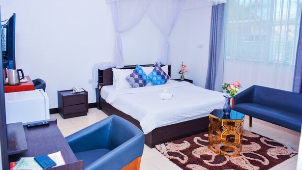 A bedroom with a bed, bedside table, nightstand, and a window with curtains. - MARKDON EXECUTIVE LODGE KISASA (Dodoma)