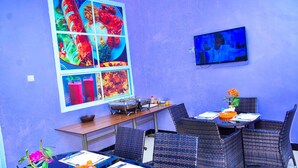 A dining area with wicker chairs, a table with a vase of flowers, and a wall-mounted TV. - MARKDON EXECUTIVE LODGE KISASA (Dodoma)