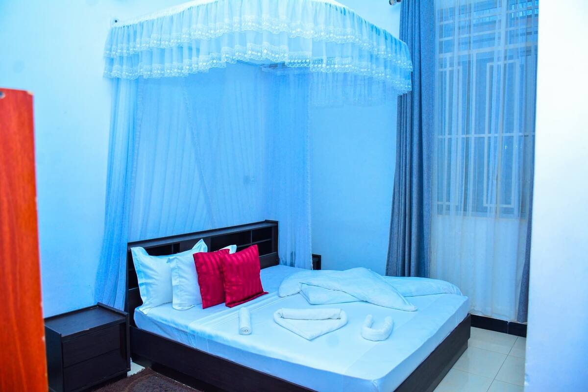 A neatly made bed with white linens and red pillows, a bedside table, and a window with sheer curtains.