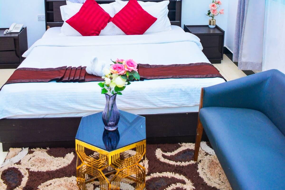 A neatly arranged bedroom with a bed, bedside tables, a bedside lamp, a vase with flowers, a blue chair, and a patterned rug.