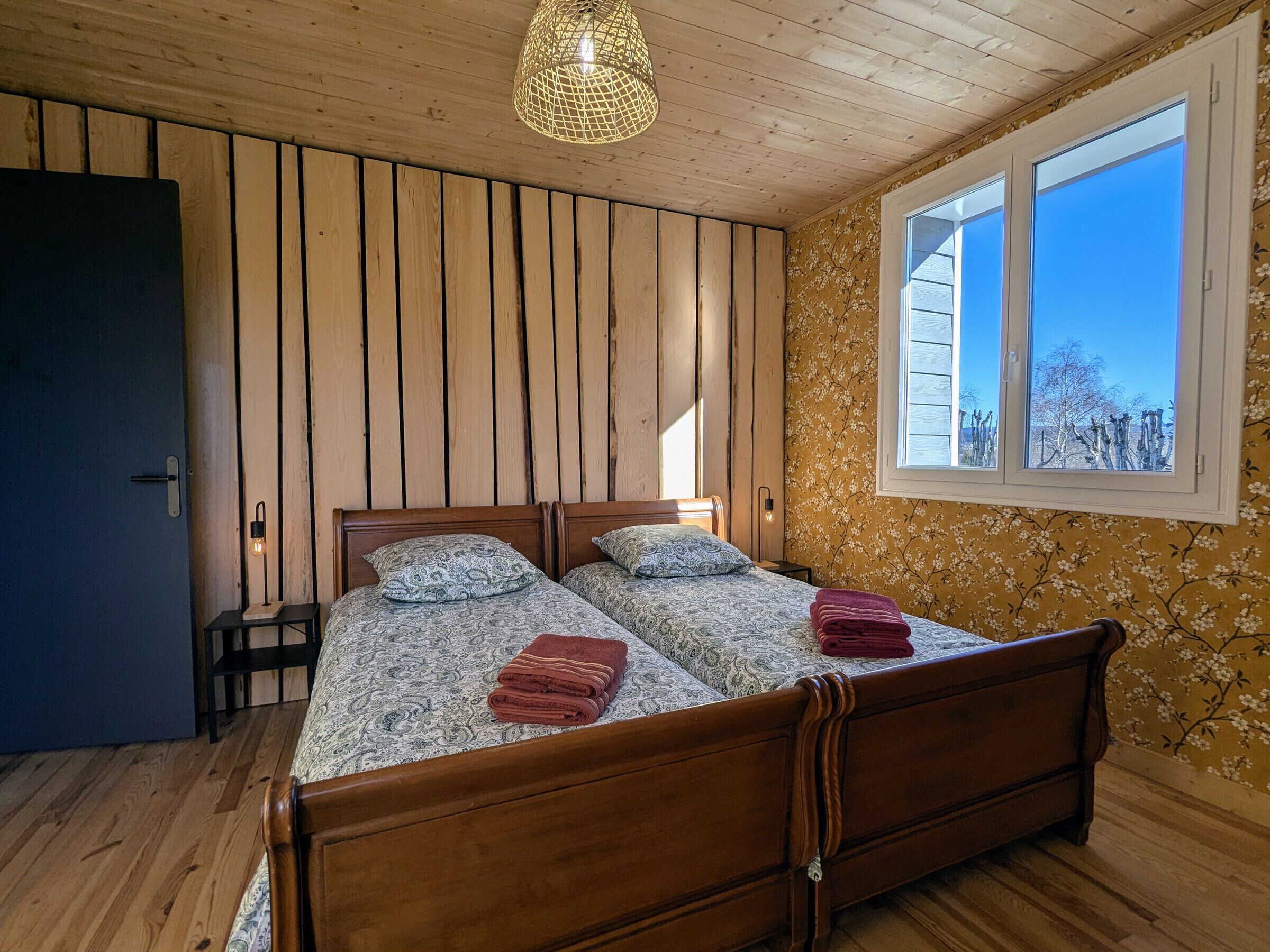 Een tweepersoonsbed met beddengoed met een patroon en twee kussens, een klein nachtkastje met een lamp, een raam met uitzicht op bomen en houten lambrisering aan de muren.
