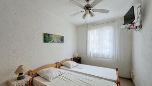 A bedroom with two single beds, a ceiling fan, a TV mounted on the wall, and a window with sheer curtains. - 2-room apartment 100m from the beach, 4 people (Argelès-sur-Mer)