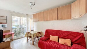 A compact living space with a red sofa, wooden dining table, and a kitchenette with wooden cabinets. - 2-room apartment 100m from the beach, 4 people (Argelès-sur-Mer)