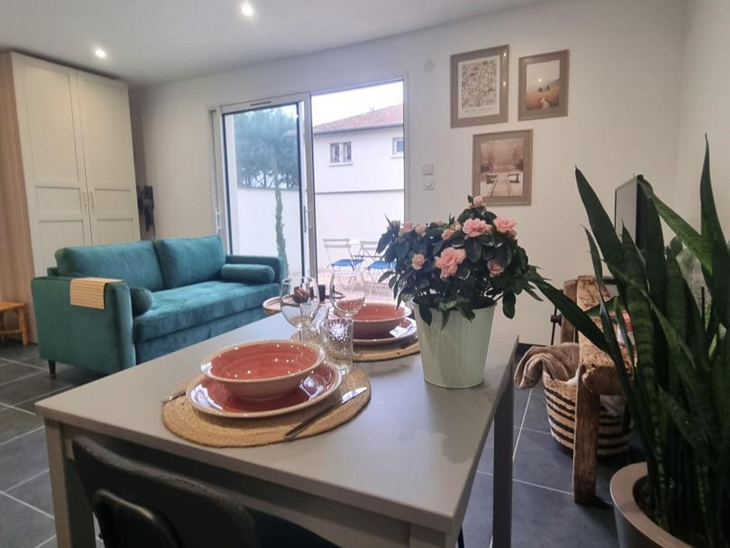 A dining area with a table set for two, a sofa, a potted plant, and a window with a view of buildings.
