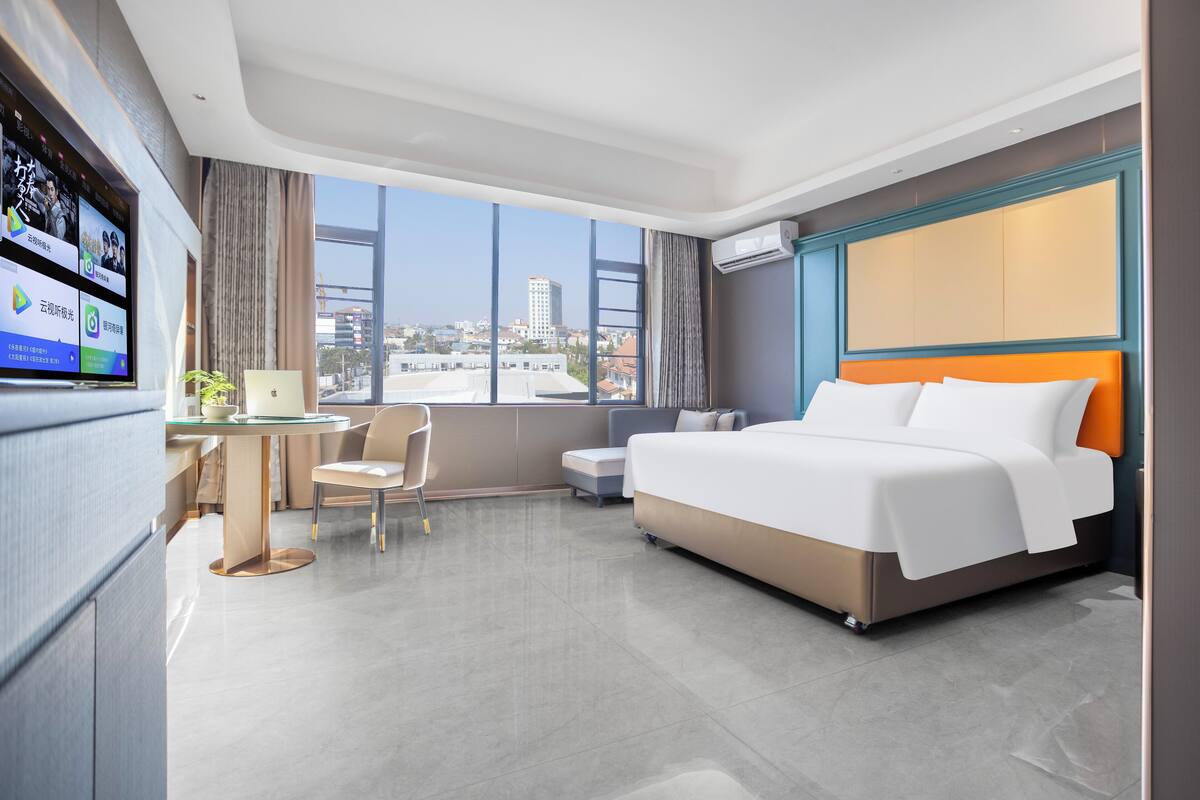 A modern hotel room with a large bed, a desk with a laptop, a chair, and a TV displaying apps.
