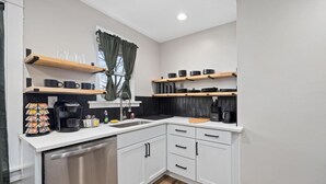 Una cocina moderna con armarios blancos, lavavajillas de acero inoxidable y encimeras negras. Hay estantes de madera sobre la encimera con varios utensilios de cocina.
