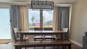 Un espace repas avec une table et des bancs en bois, un luminaire au plafond et une vue sur l’extérieur par une fenêtre agrémentée de rideaux.