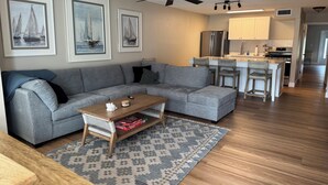 Un salon moderne avec un canapé d’angle gris, une table basse en bois et un tapis à motifs. On peut voir des œuvres d’art encadrées au mur, un ventilateur de plafond et, au fond, un coin cuisine.