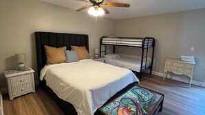 Une chambre avec un grand lit, un lit superposé plus petit, un ventilateur de plafond, une table de chevet avec une lampe et une œuvre d’art colorée posée sur le sol.