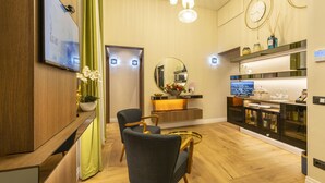 Una habitación de hotel moderna con piso de madera, un televisor de pantalla plana montado en la pared, un espejo redondo sobre el mostrador y una zona de estar con dos sillas y una mesa de centro de vidrio.