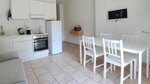 A modern kitchen with white cabinets, a built-in oven, and a dining area with white chairs and a table. - Dinky's apartment with forest view balcony (Magreglio)