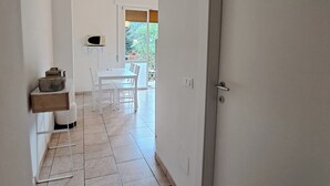 A narrow hallway with a white door on the right, a white table and chairs in the background, and a small shelf with items on the left. - Dinky's apartment with forest view balcony (Magreglio)