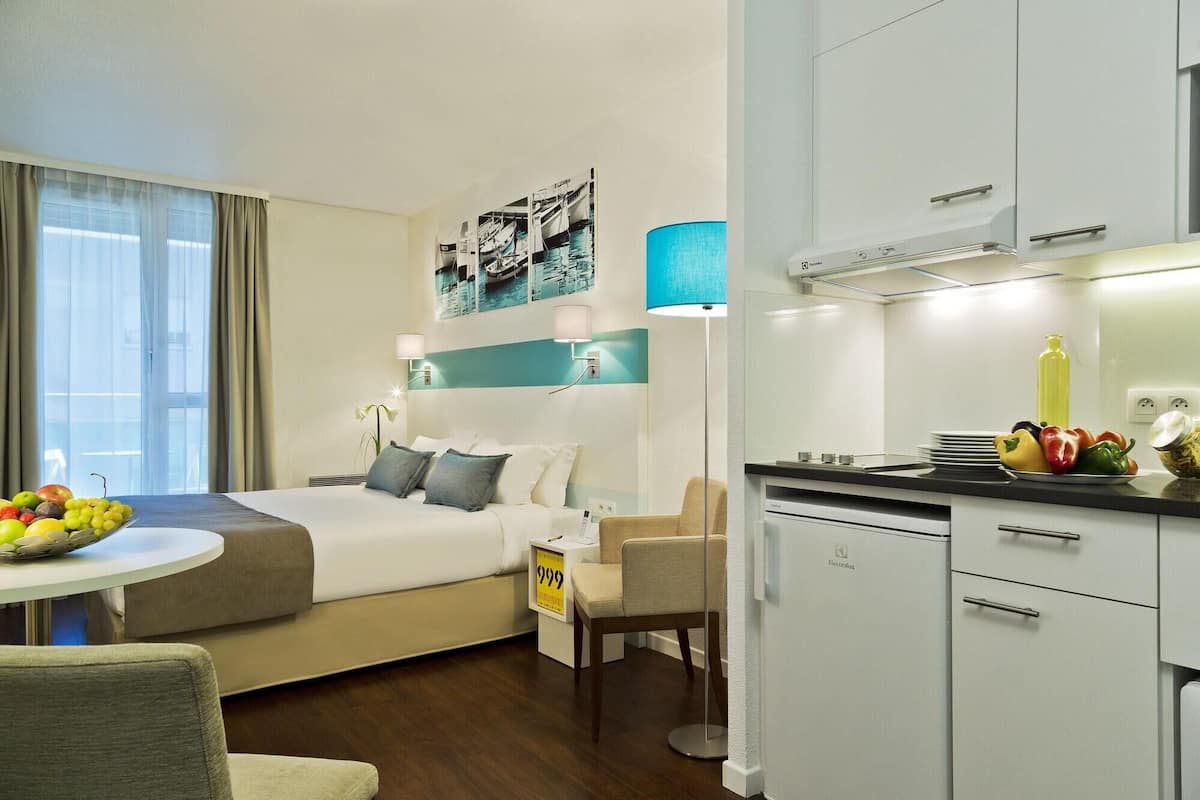 Une chambre d’hôtel comprenant un lit, une table de chevet avec des fruits, une chaise, un bureau et une kitchenette.