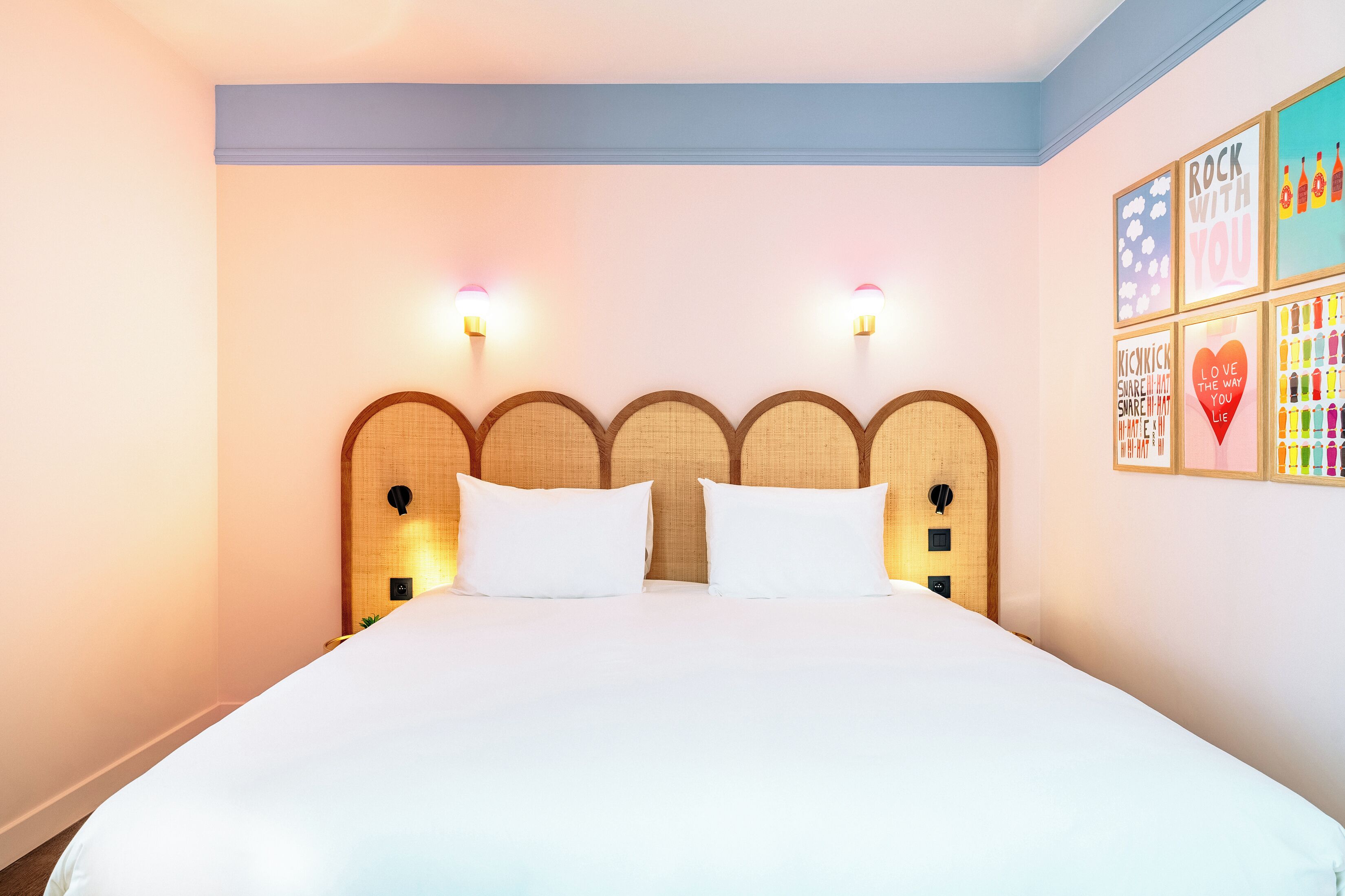 A hotel room with a large bed, two wall-mounted lights, a headboard with a woven design, and a wall with colorful framed artwork.