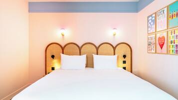 A hotel room with a large bed, two wall-mounted lights, a headboard with a woven design, and a wall with colorful framed artwork.