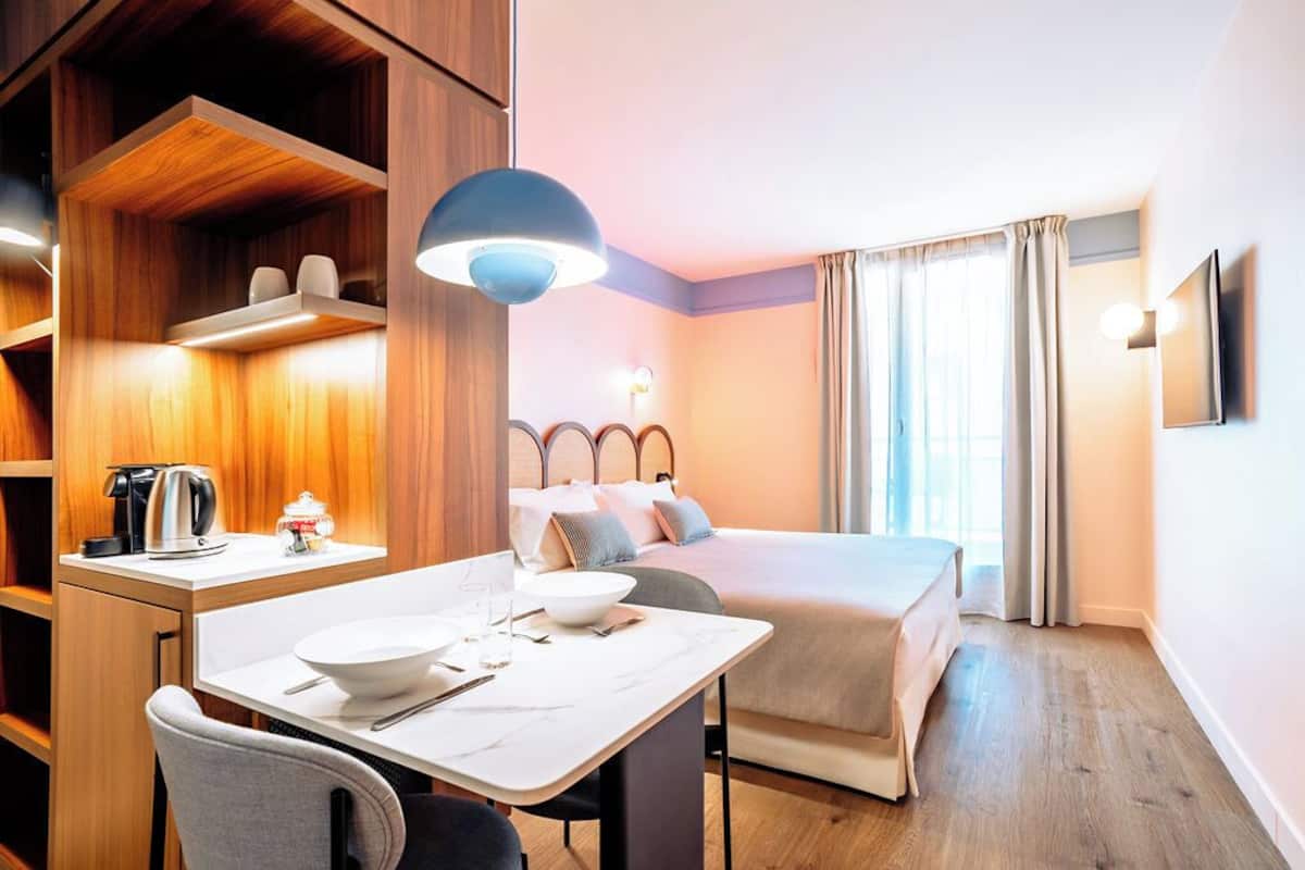Une chambre d’hôtel avec un lit, une table de chevet et un bureau avec une chaise. Il y a une fenêtre avec des rideaux, un téléviseur fixé au mur et un luminaire au plafond.