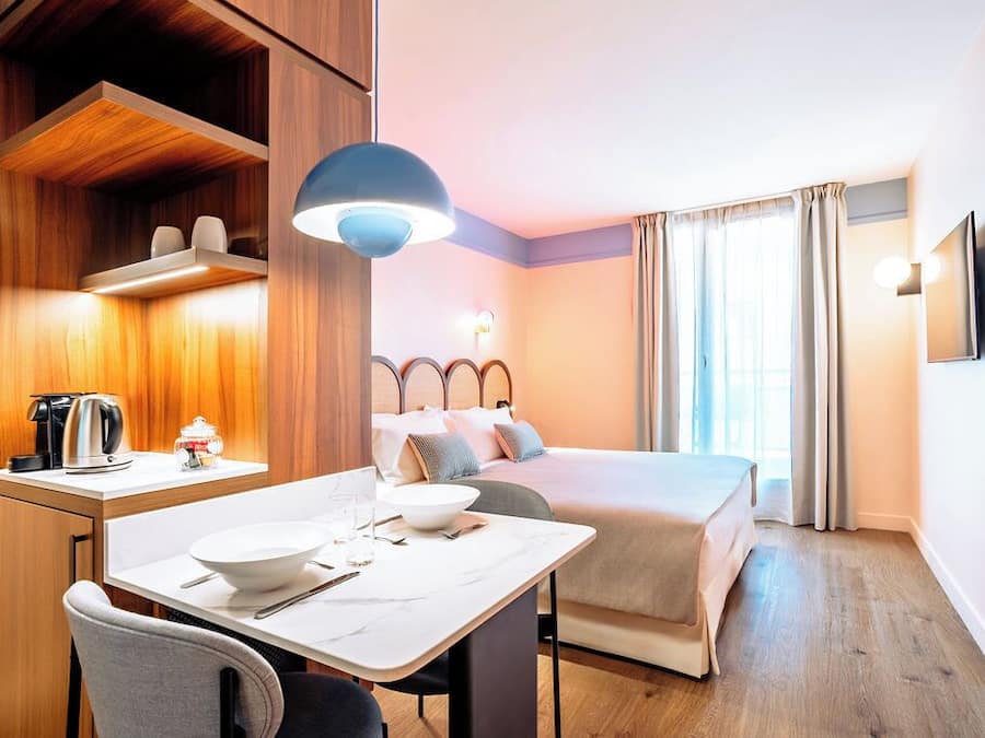 A hotel room with a bed, bedside table, and a desk with a chair. There is a window with curtains, a wall-mounted TV, and a ceiling light.