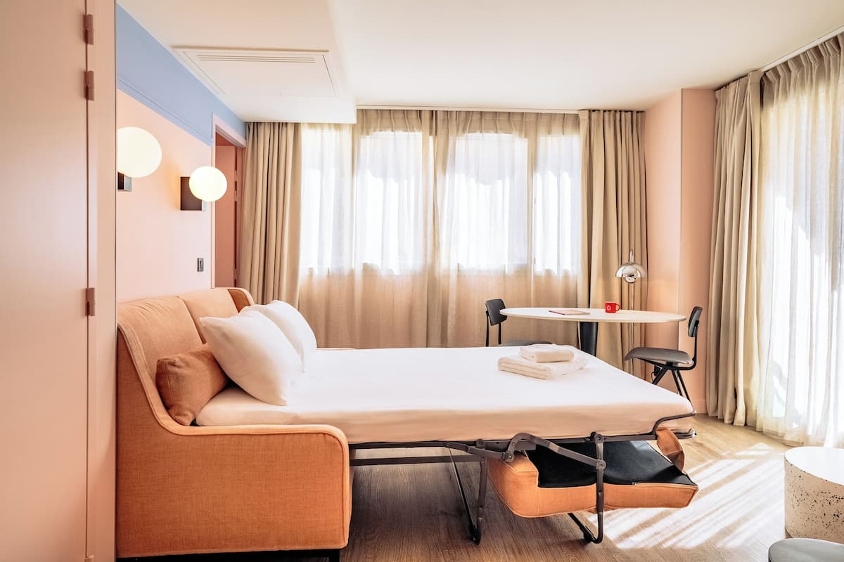 Une chambre d’hôtel comprenant un lit, une petite table avec une chaise, une table ronde avec une chaise, et une grande fenêtre avec des rideaux.