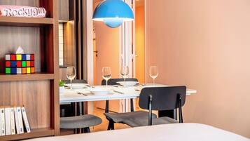 A modern dining area with a white table set for two, a blue pendant light, and a bookshelf with a Rubik's Cube and books.