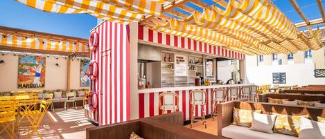 A bar area with striped awnings, a seating area with wooden benches and cushions, and a bar counter with shelves of bottles.