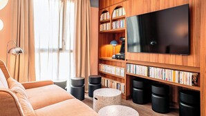 Een moderne woonkamer met een grote flatscreen-tv, houten boekenplanken, een bank en ronde, zwarte krukken.