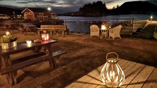 Een terras buiten met houten banken en tafels, een lantaarn en uitzicht op een meer in de schemering.