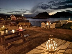 An outdoor seating area with wooden benches and tables, a lantern, and a view of a lake at dusk. - Fantastic fishing hut with sea view directly on the water (Korshamn)
