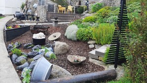 Een tuin met verschillende planten, stenen en een zwarte metalen sculptuur.
