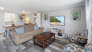 A modern living room with a sofa, coffee table, and TV. There is a kitchen area with a dining table and chairs.