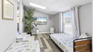 A room with two beds, a large window with curtains, a ceiling fan, a potted plant, and a painting on the wall.