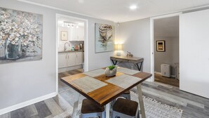 A modern dining area with a wooden table, cushioned stools, and a central plant. A kitchen is visible in the background with white cabinets and a countertop.