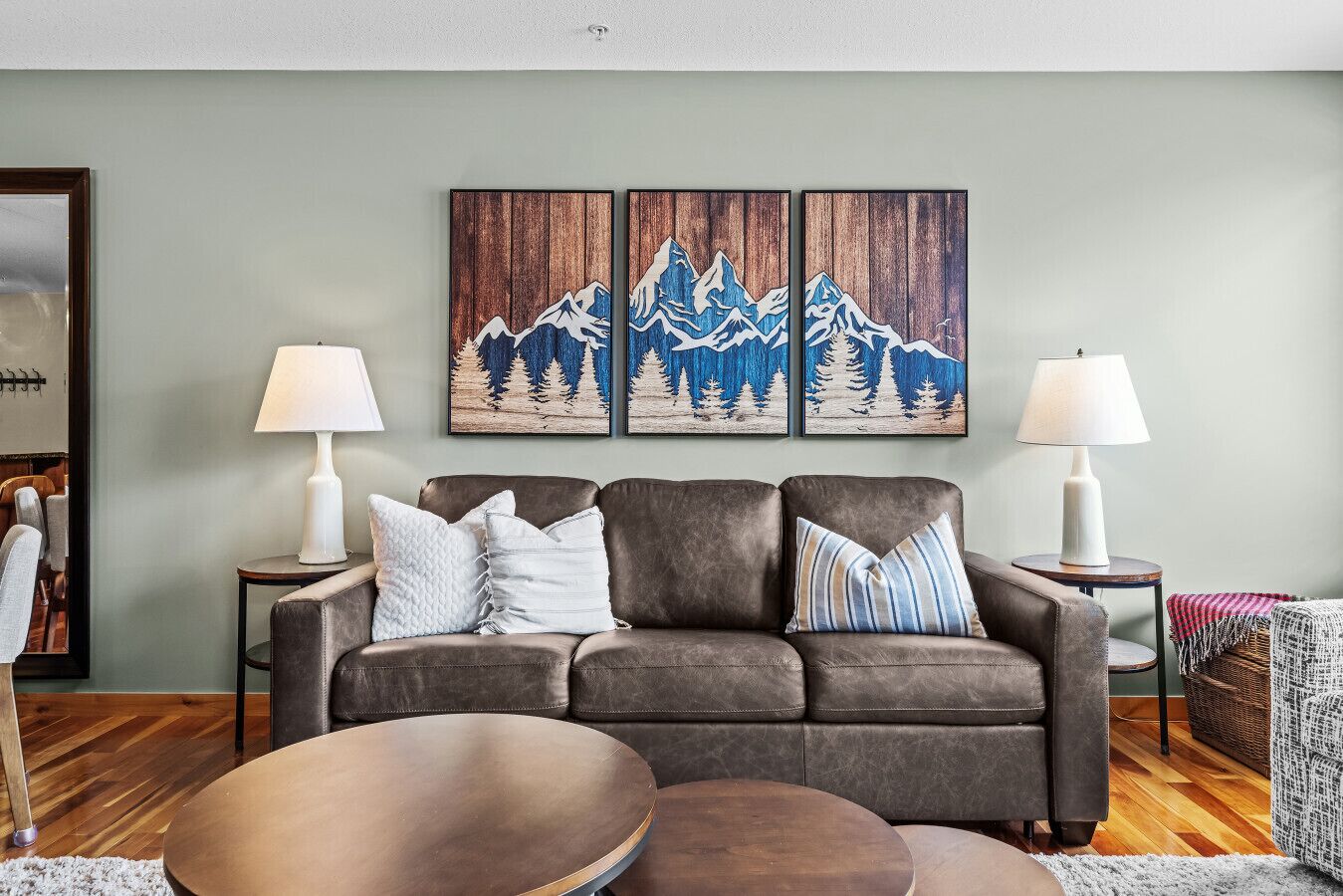 A living room with a brown leather sofa, two side tables with lamps, a round coffee table, and a large painting of mountains on the wall.
