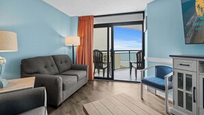 Une piÚce avec un canapé gris, une petite table avec une lampe, une chaise bleue et un meuble. Il y a un balcon avec vue sur la plage.