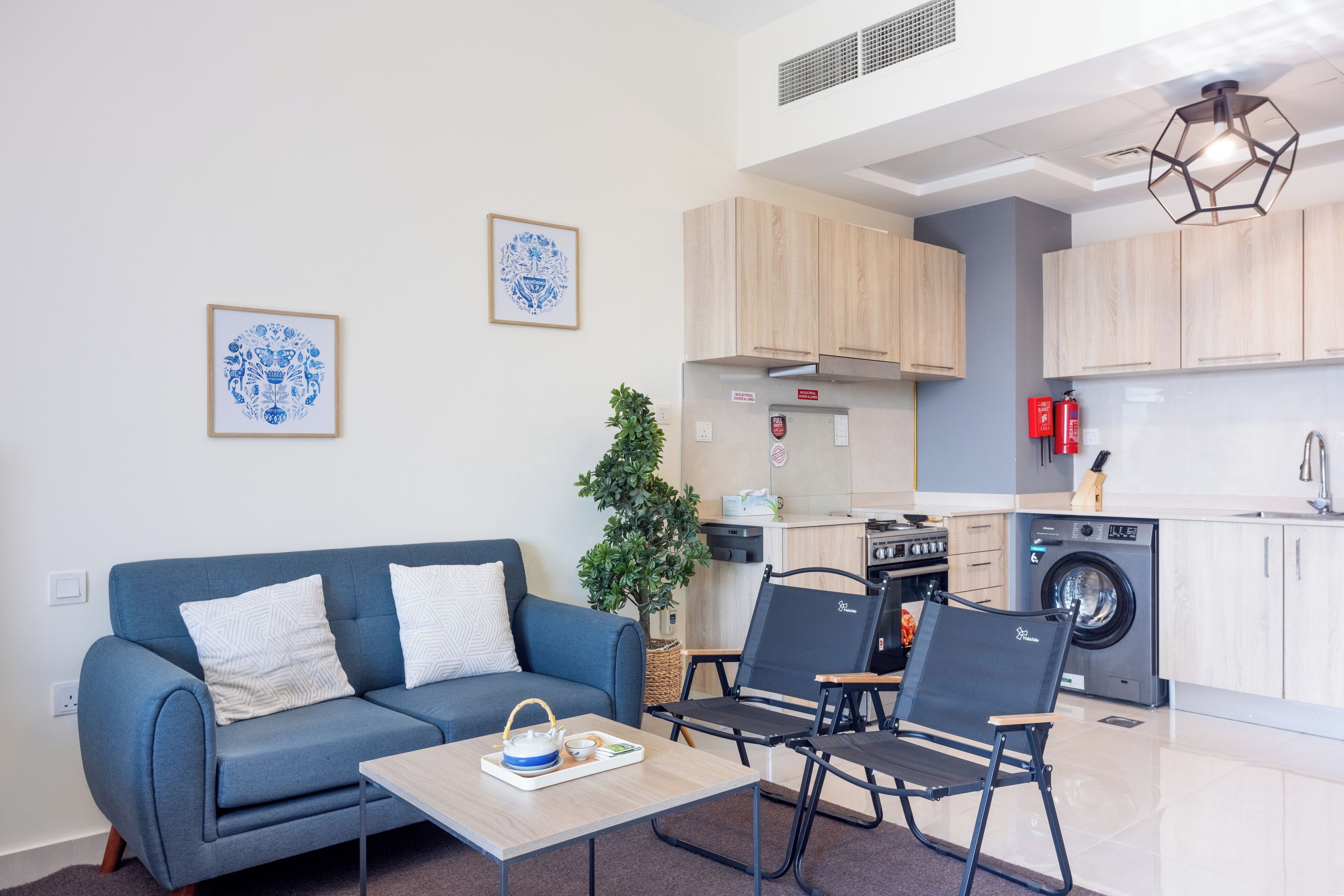 Een modern appartement met een blauwe bank, een eettafel, een keuken met houten kastjes, een wasmachine en een plafondlamp.