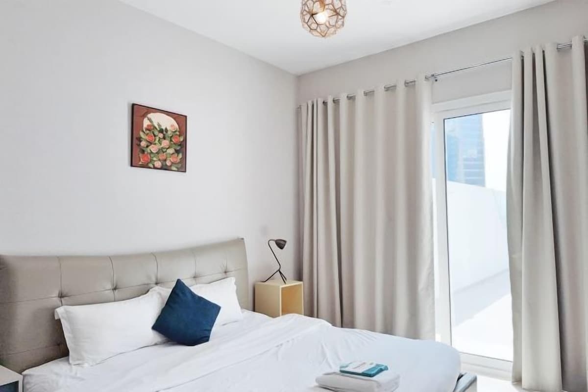 A hotel room with a bed, a bedside table, a lamp, and a window with curtains.