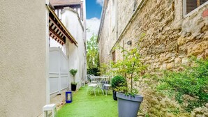 Un patio estrecho con césped artificial verde, sillas de metal blanco y plantas en macetas.