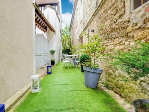 A narrow courtyard with artificial grass, a white metal table and chairs, potted plants, and a stone wall. - Romantic Stay, Isigny-sur-Mer (Isigny-sur-Mer)