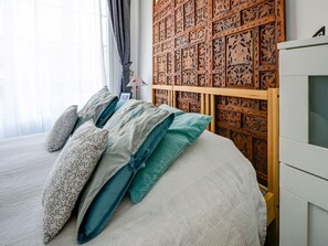 Una cama bien tendida con almohadones estampados, un cabecero de madera con intrincados grabados y una mesita de noche blanca a la derecha.