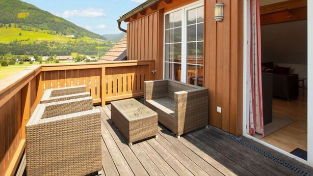 Una terraza de madera con mobiliario de mimbre, un ventanal de vidrio que da al interior y vistas a colinas verdes.