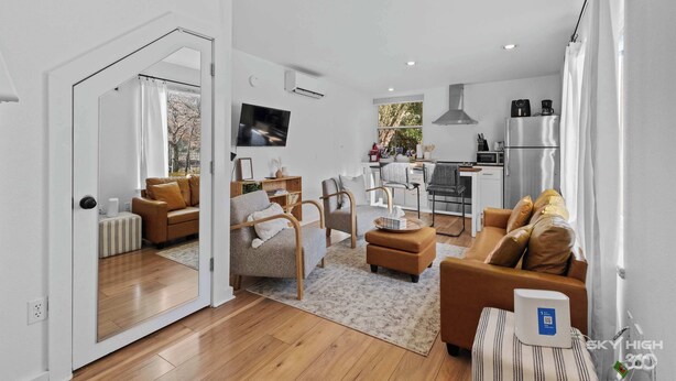 A modern living room with a sofa, armchairs, and a coffee table. There is a kitchen area with a refrigerator, stove, and microwave. A TV is mounted on the wall, and there is a window with curtains.