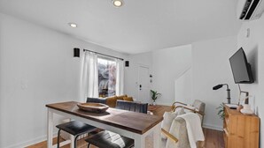 A modern, well-lit room with a wooden dining table, two black chairs, a sofa with a white throw, and a TV mounted on the wall.