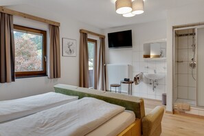 A bedroom with two beds, a TV, a bathroom with a shower, and a window with curtains.