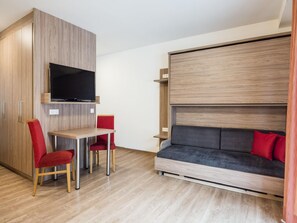 Une pièce de taille modeste, avec un téléviseur fixé à un mur en bois, une petite table accompagnée de deux chaises et un canapé agrémenté de coussins rouges.