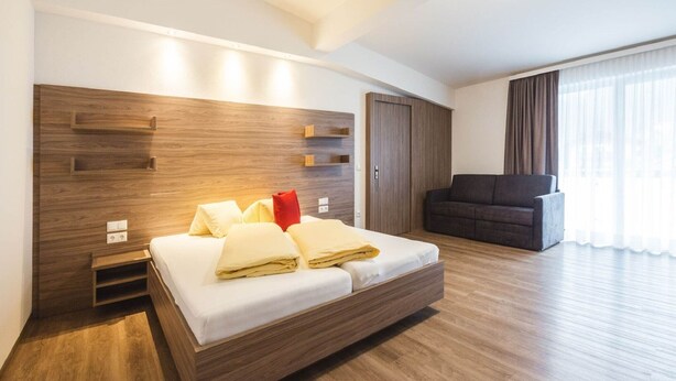 Une chambre d’hôtel moderne avec une tête de lit en bois, un lit recouvert de draps blancs, un canapé foncé et une grande fenêtre agrémentée de rideaux légers.